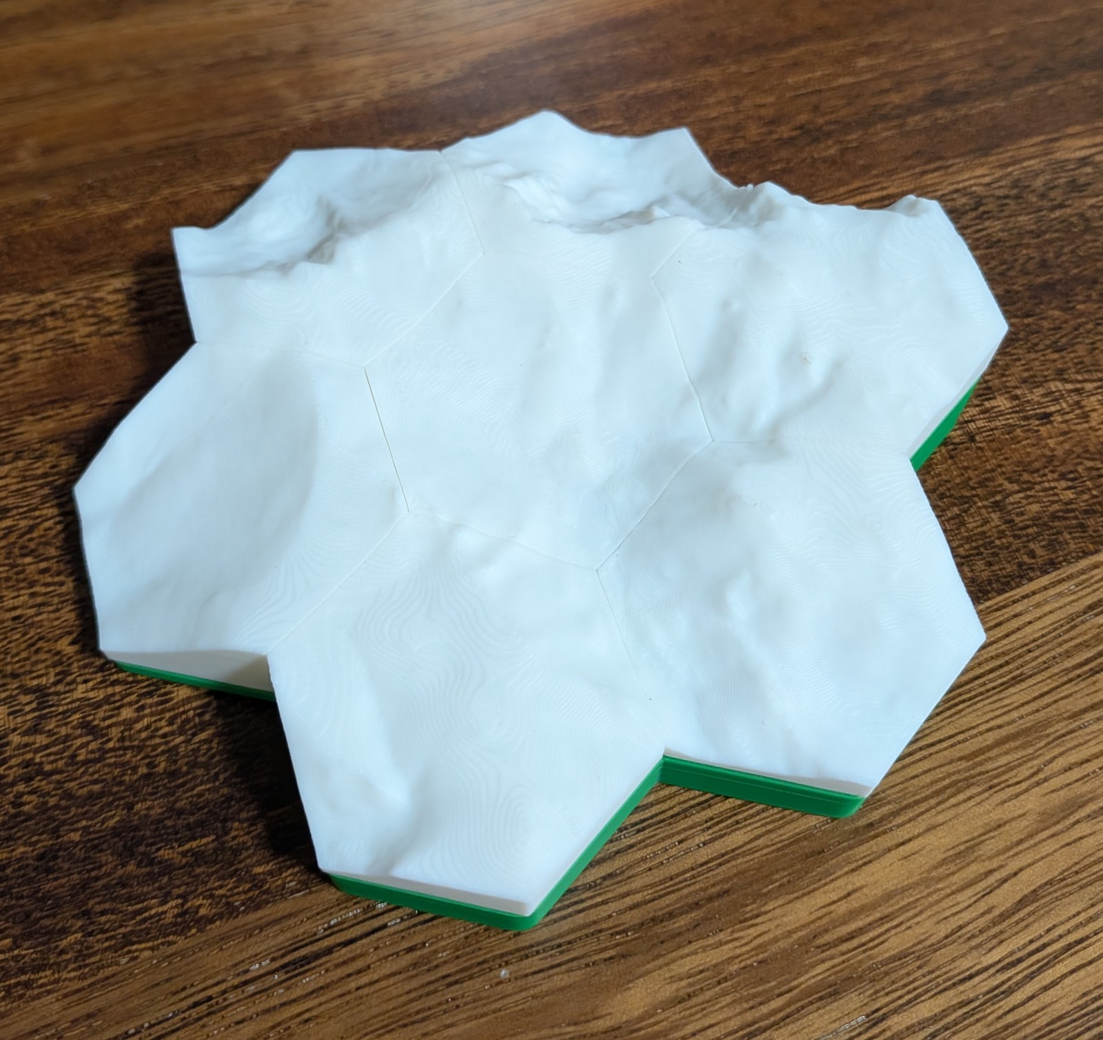 Seven printed hexagonal terrain tiles assembled on a green pedestal, showing continuous mountain terrain across the seams