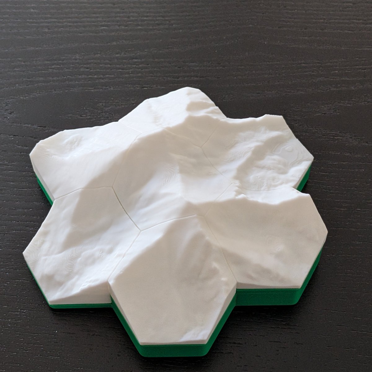 Seven-tile topographic set with white peaks over a green base layer, angled on dark wood